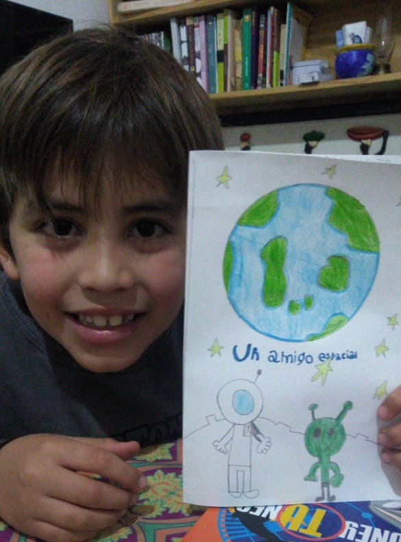 Niño mostrando una hoja que dice: Mi amigo espacial con el dibujo de un niño en la luna y el de un marciano en color verde